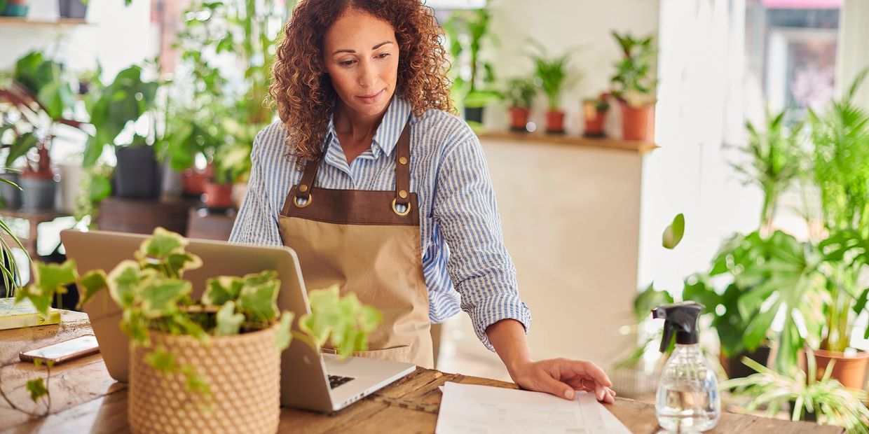 A woman in an apron working with a laptop and plants around her.