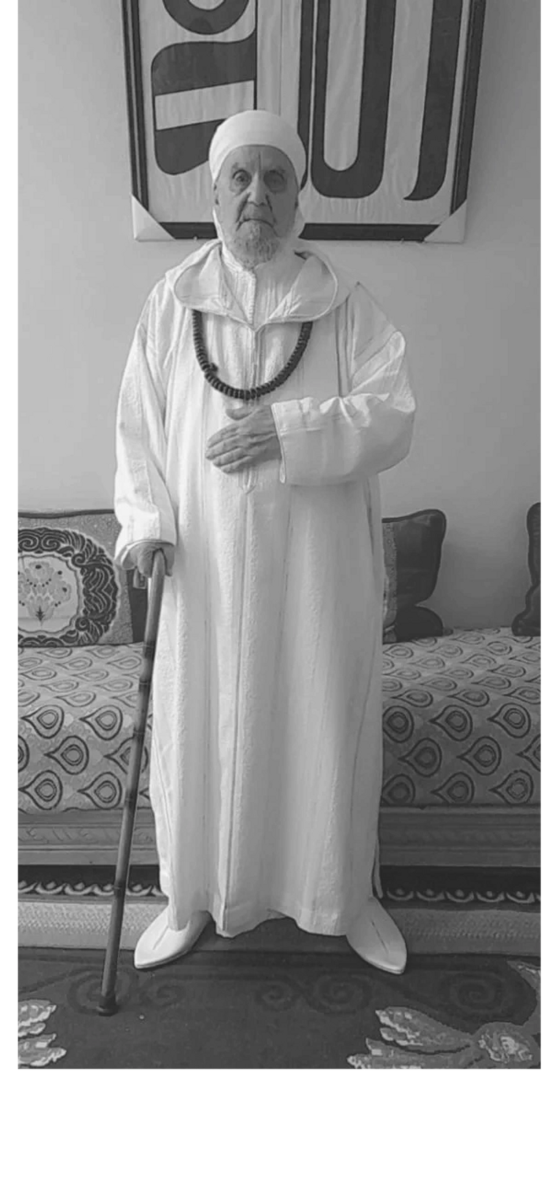Elderly man in traditional white robe with a walking cane, standing indoors.