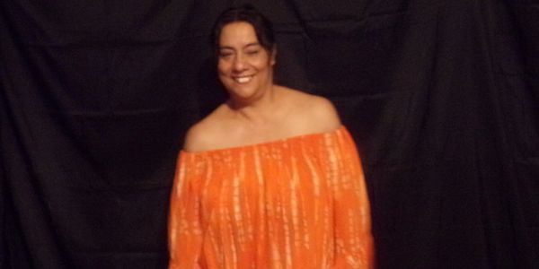 A smiling woman in an orange off-shoulder dress against a black backdrop.