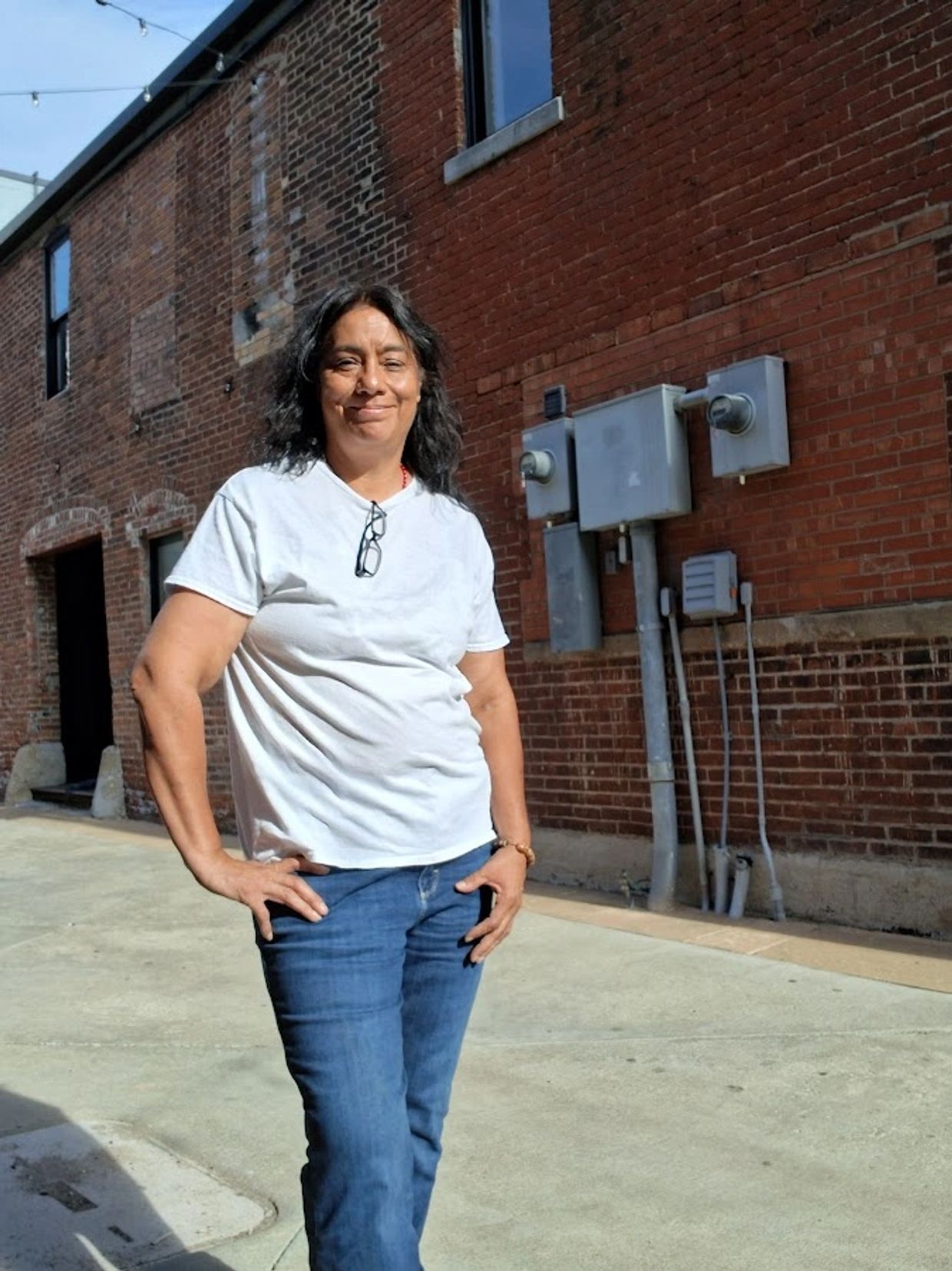 A woman in a white t-shirt and jeans stands smiling by a brick wall.