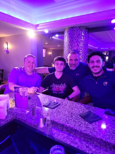 Four men smiling at a bar under purple lighting.
