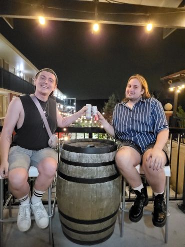 Two friends toasting drinks over a barrel table at night.