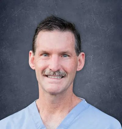 Smiling man in light blue scrubs against a dark background.