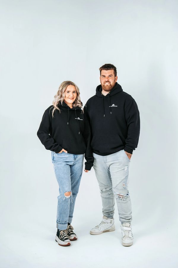 Two people in black hoodies and ripped jeans standing against a white background.