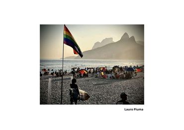 Rainbow flag waves on a crowded beach at sunset with mountainous backdrop.