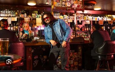 Man in denim jacket posing confidently in a lively bar setting.