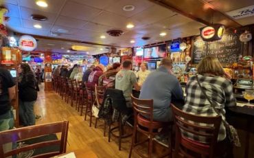People sit at a lively bar with wooden floors and warm lighting.