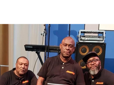 Three men in black polo shirts posing indoors with musical equipment.