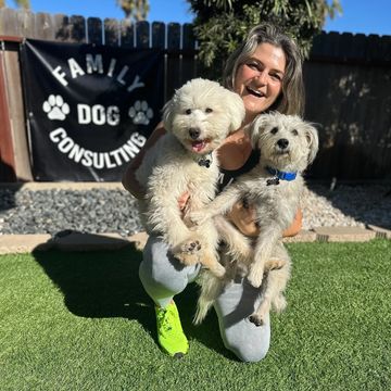 Happy woman holding two small dogs in front of a Family Dog Consulting banner.