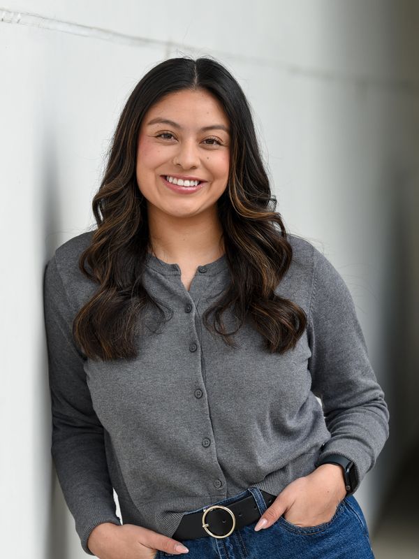 Woman with long dark hair smiling, wearing a gray cardigan and jeans, leaning against a white wall.