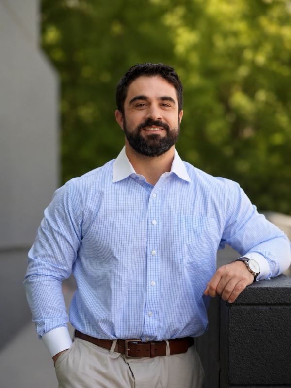 Smiling man in blue checkered shirt leaning on a wall outdoors.