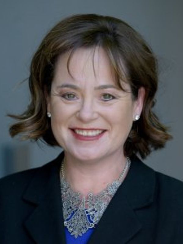 Smiling woman with short brown hair wearing a black blazer and statement necklace.