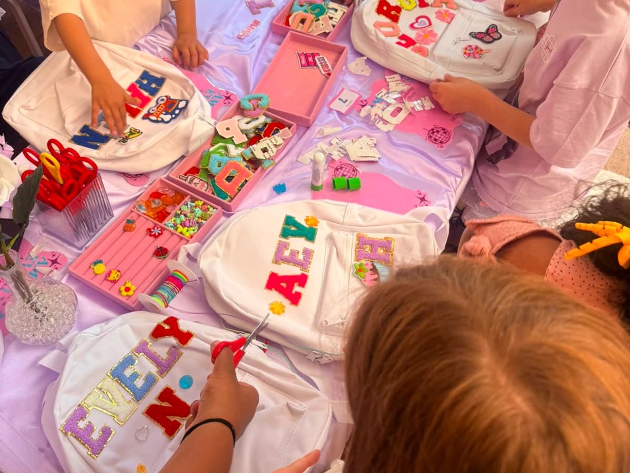 Kids decorating white backpacks with colorful patches and letters.