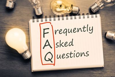 Notebook with FAQ acronym surrounded by light bulbs on a wooden table.