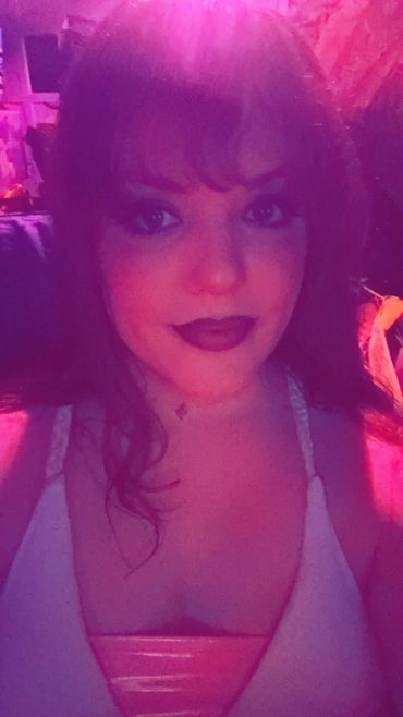 In low pink lighting, a sweet faced t-girl has on a white dress and dark lipstick.