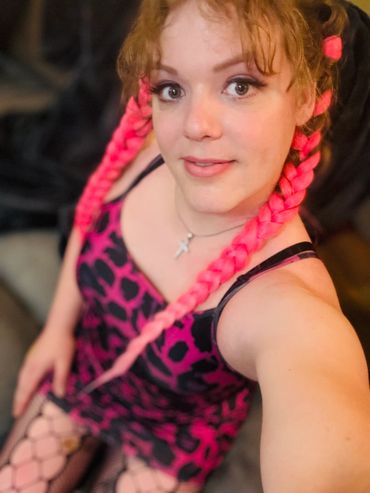 Spunky MTF TS girl sits and smiles at camera. Her hair is braided and pink, her dress is pink leopard