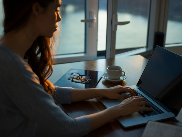 A woman working on a laptop near a window with a cup and glasses on the table.