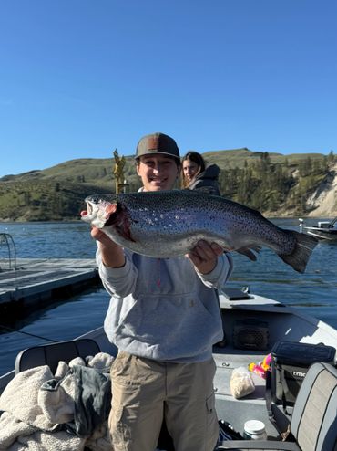 A young man proudly holds a large trout on a boat with a scenic lake and hills behind.