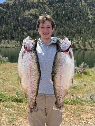 Young man holding two large fish with a scenic lake and forest background.