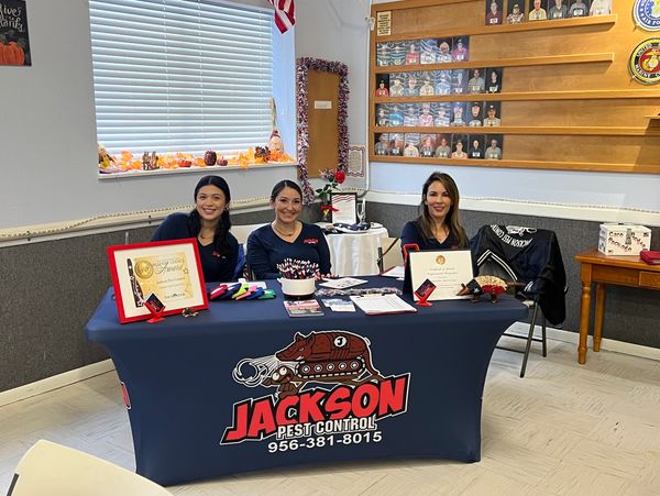 Three women at a Jackson Pest Control promotional table with awards and brochures.