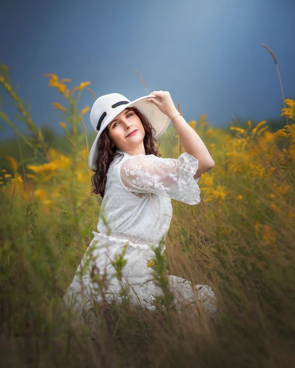 a woman in a white vintage dress kneeling in grass while holding her hat
