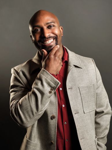 Smiling man in beige coat and red shirt, thoughtfully touching his chin.