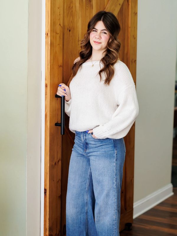 Young woman in white sweater and blue jeans standing by a wooden door.
