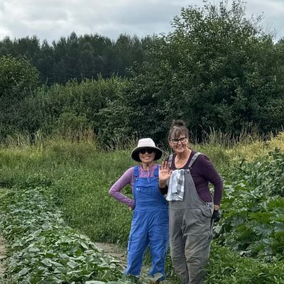 Two women in overalls standing in a lush green garden, smiling and waving.
