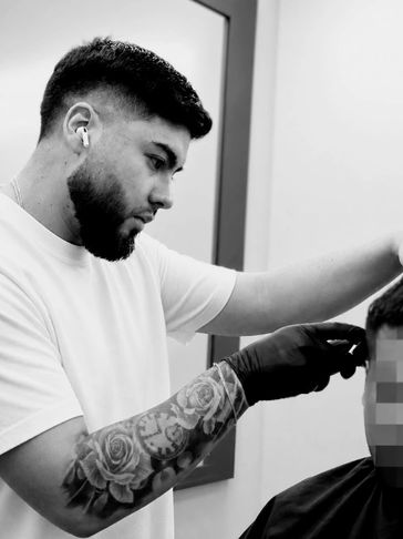 Barber with tattooed arm cutting a client's hair wearing a white shirt.