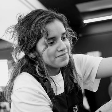 Young woman with wavy hair focused on her work, wearing a white shirt and apron.