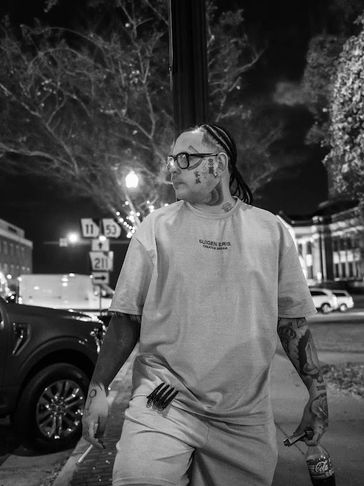 A tattooed man with braided hair and glasses stands on a city street at night.