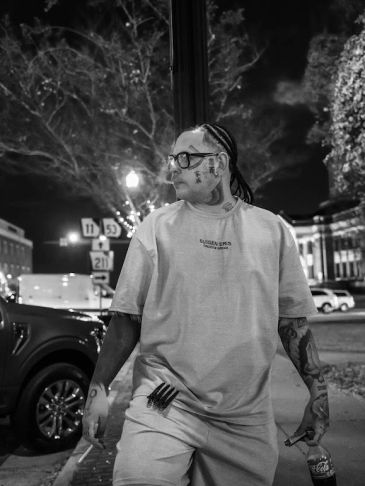 A tattooed man with braided hair and glasses stands on a city street at night.