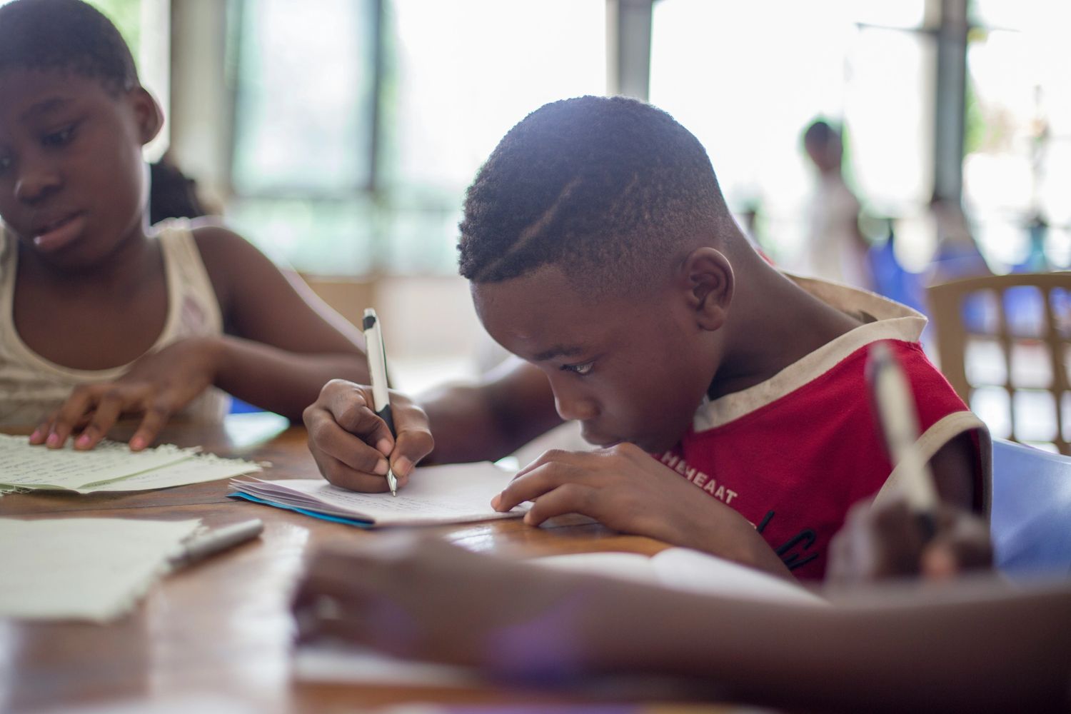 Children focused on writing and studying at a table in a classroom.