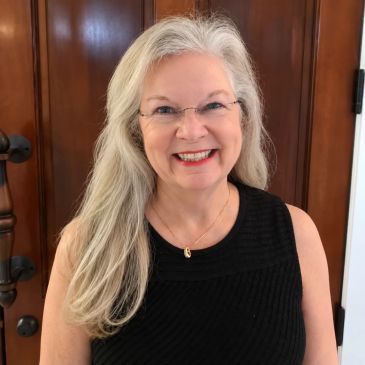 Smiling woman with long gray hair and glasses in black top.