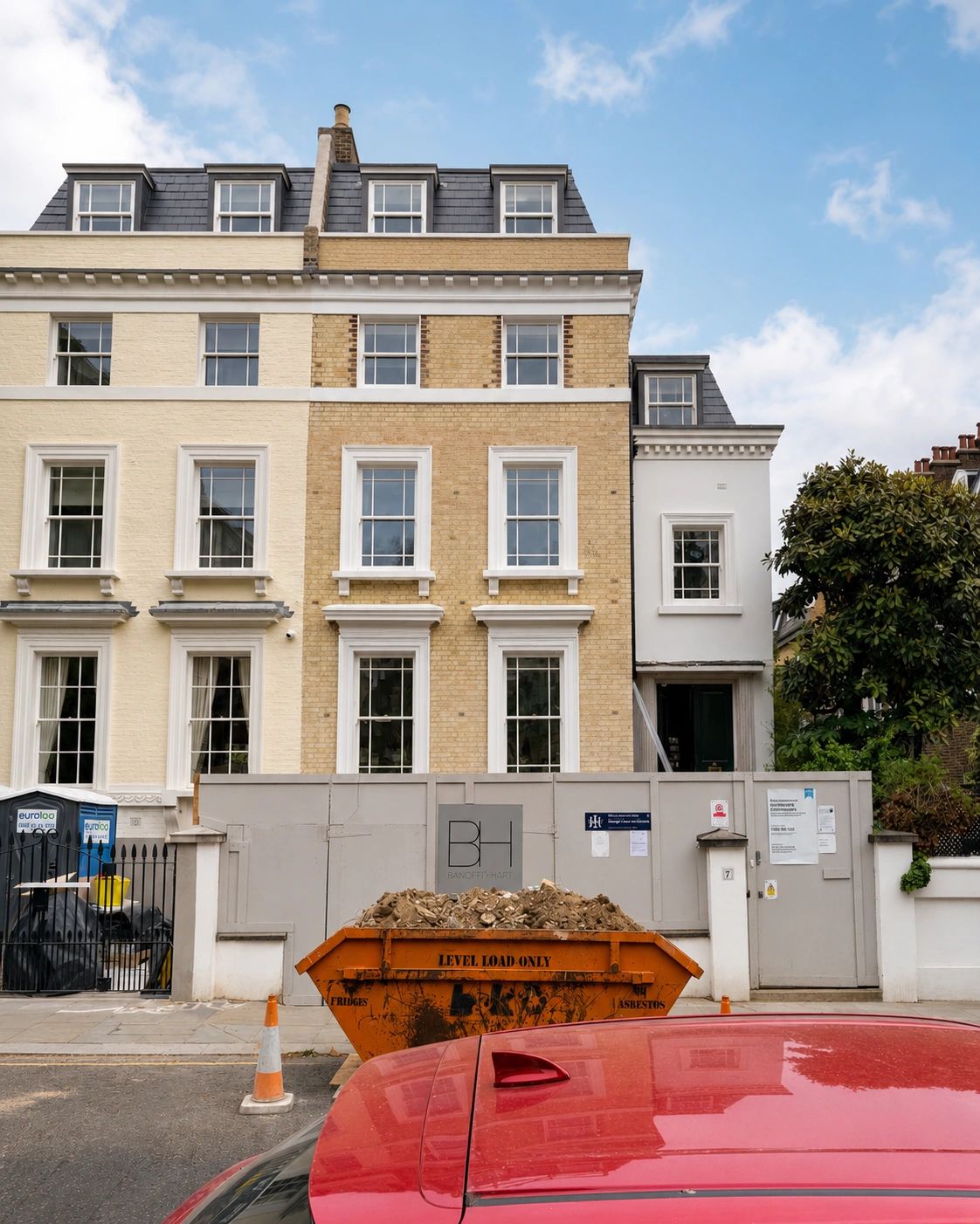 Construction site with a skip bin and a red car in front of a renovated historic building.