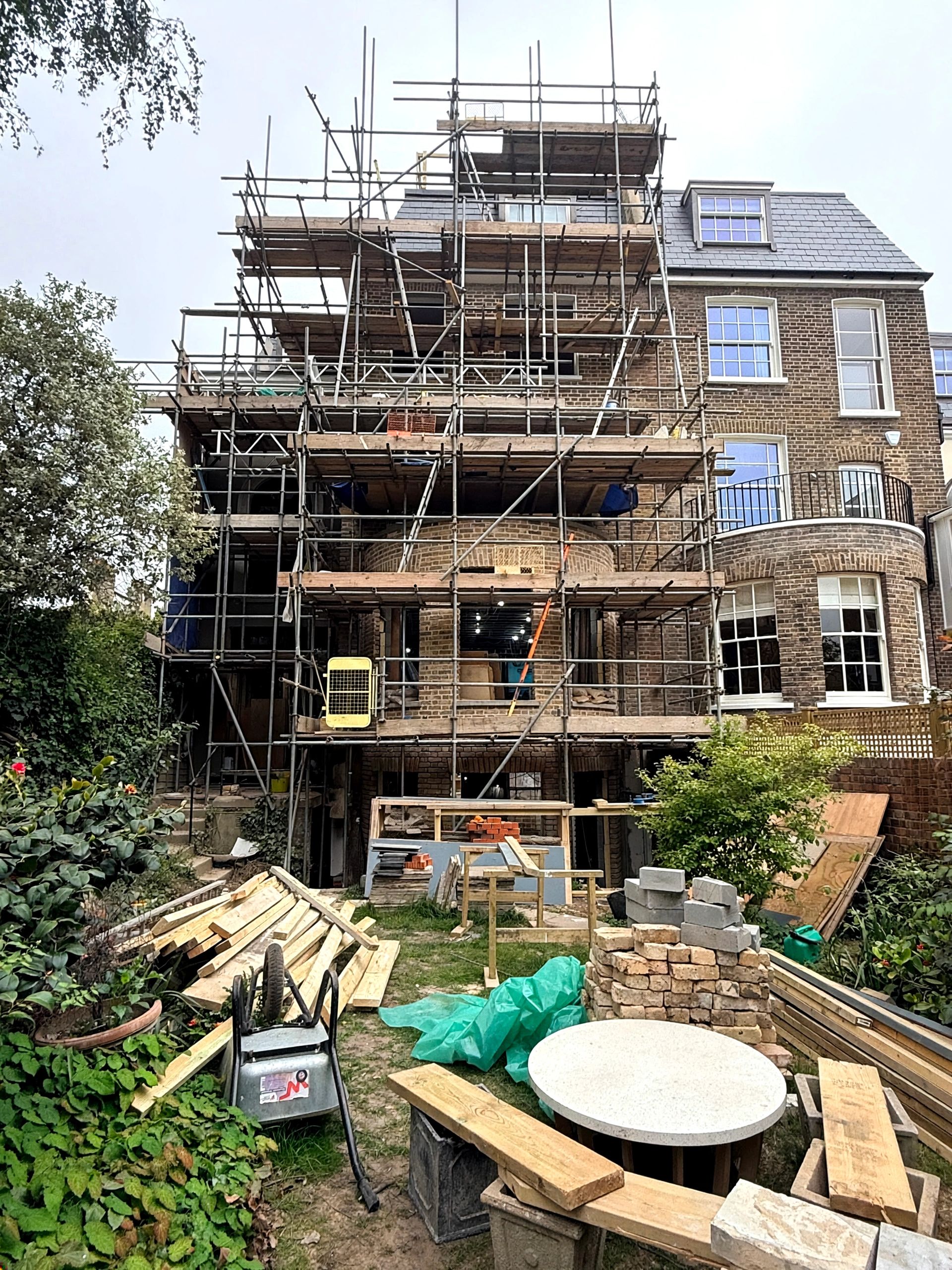 A brick house under renovation with scaffolding and construction materials in the garden.