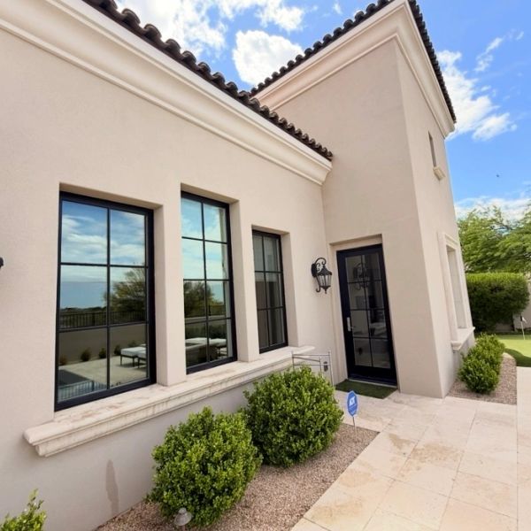 Modern beige house exterior with black-framed windows and lush green bushes.