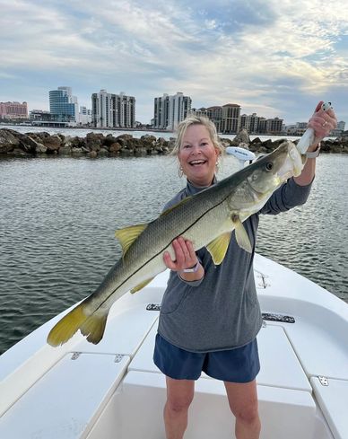 Woman proudly holding a large fish on a boat with city buildings in the background.