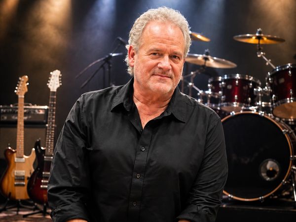 A mature man with curly gray hair in a black shirt stands in front of musical instruments on stage.