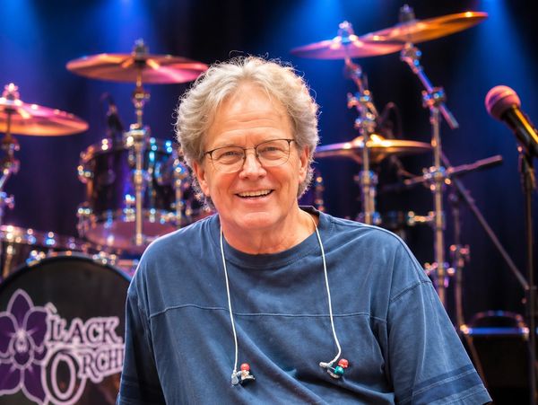 A smiling man with glasses sits in front of a drum set on stage.