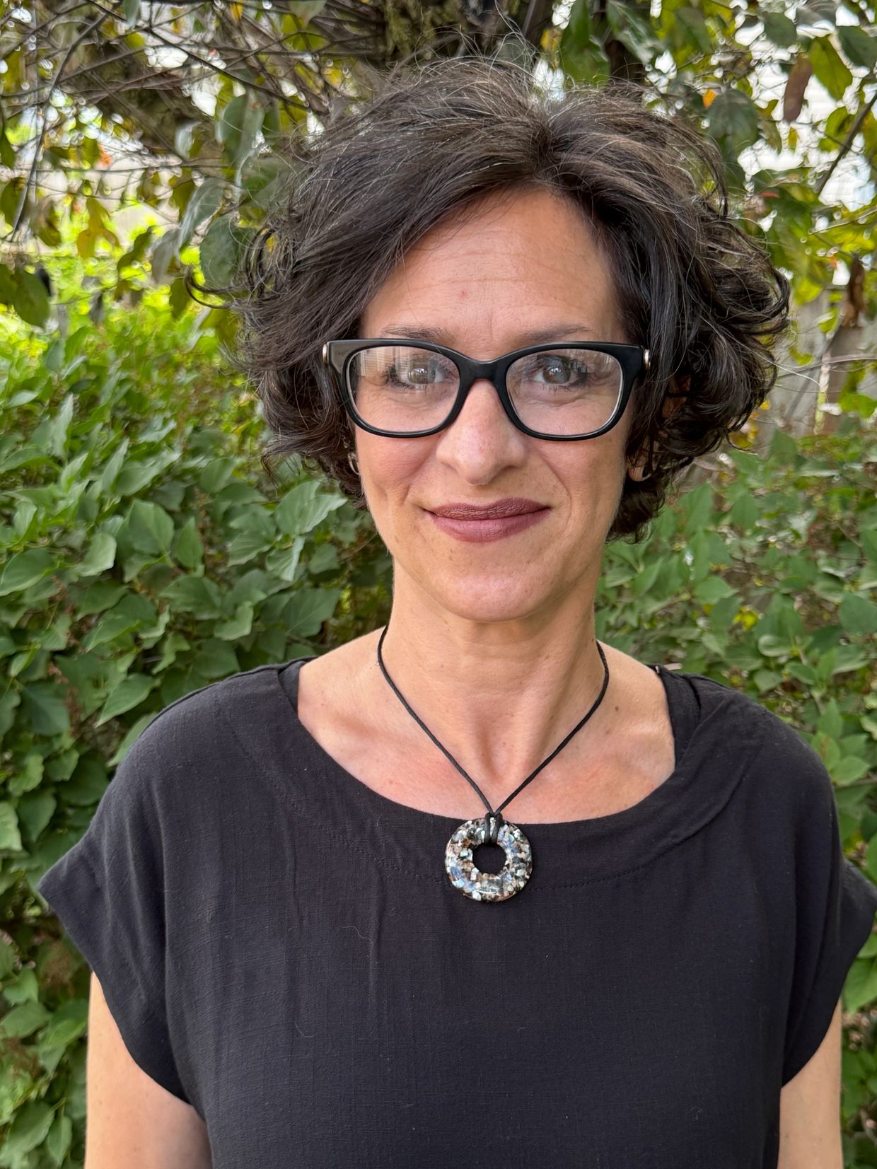 Smiling woman with short curly hair and glasses stands outdoors.