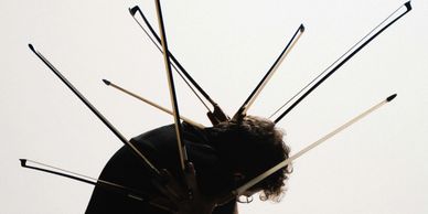 Person holding multiple violin bows radiating from their back against a light background.
