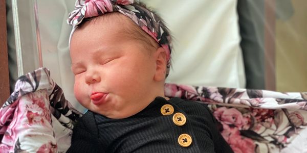 Newborn baby sticking out tongue, dressed in black, lying on floral blanket.