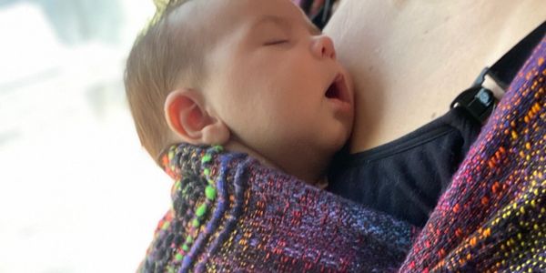 A baby peacefully sleeping in a colorful woven wrap on someone's chest.