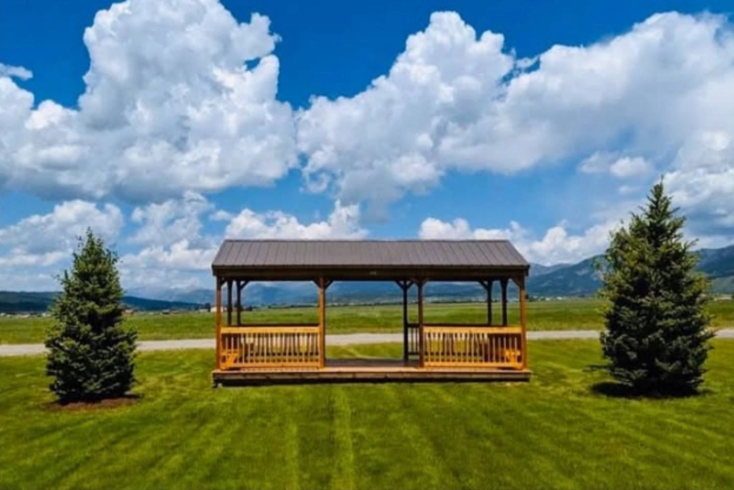 Open wooden gazebo with benches between two pine trees on a grassy field.