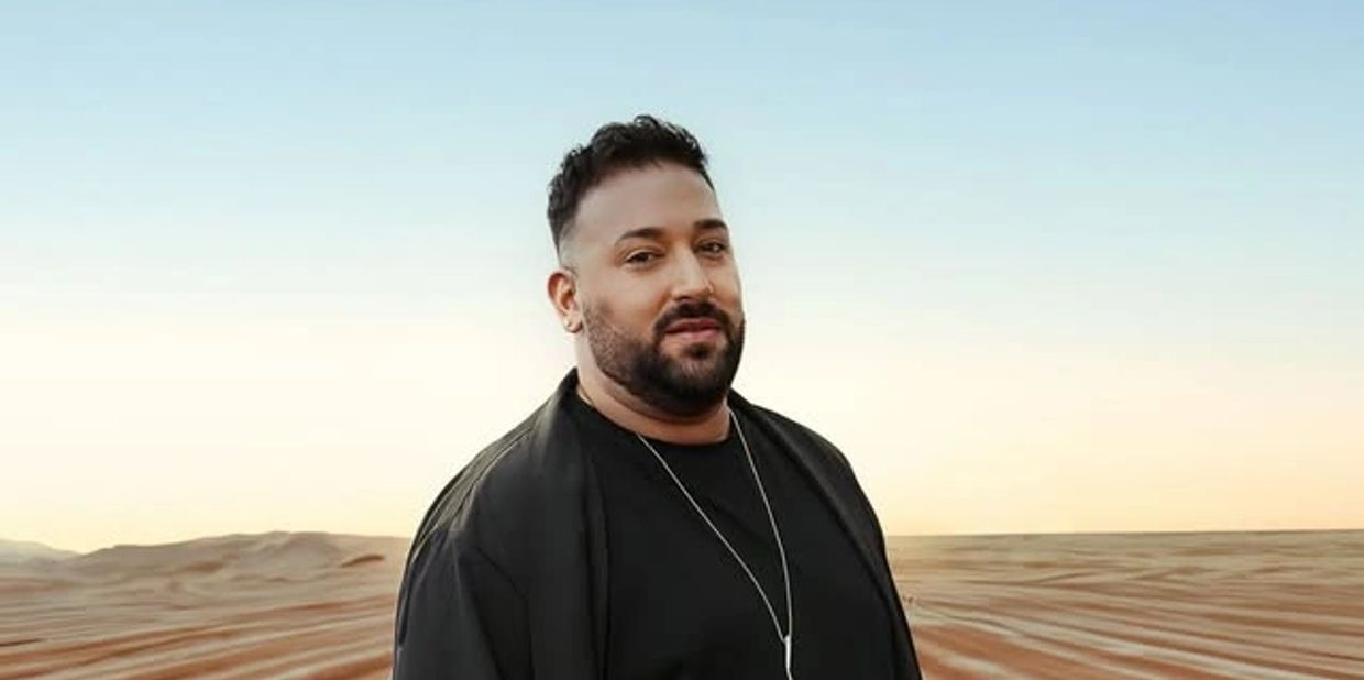 Man with a beard in a black outfit standing in a desert.