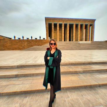 A woman in a black and green robe stands confidently on stone steps in front of a grand building.