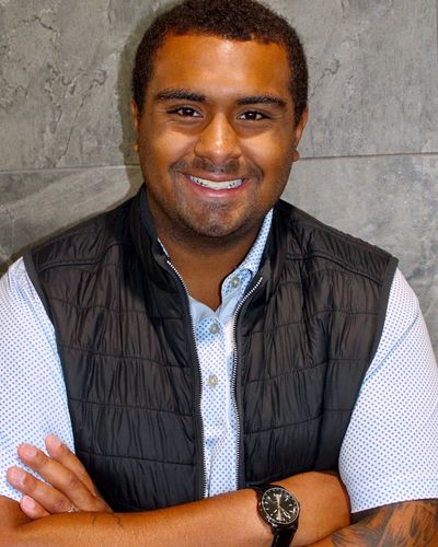A smiling young man with crossed arms wearing a black vest and watch.
