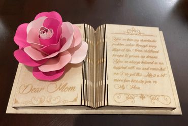 A wooden book-shaped card with a pink paper flower and a message for Mom.