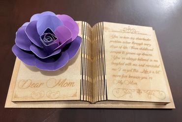 A wooden book-shaped plaque with a purple paper flower and a heartfelt message for Mom.